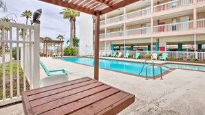 A pool area with lounge chairs and a wooden table, surrounded by a multi-story building with balconies. - Oceanfront Corpus Christi Condo w/ Pool! (Corpus Christi)