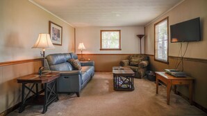 A living room with a blue sofa, a wooden coffee table, a patterned armchair, a lamp, a TV, and a window with blinds. - Lakefront Cabin in Dandridge w/ Fire Pit & Grill! (Dandridge)