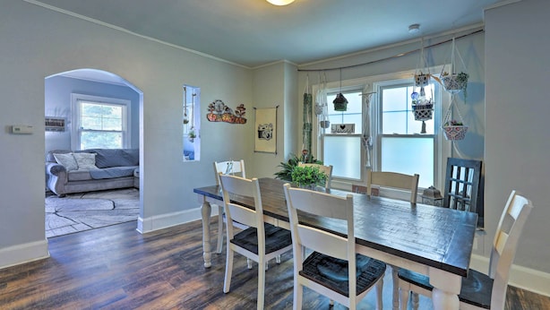A dining area with a wooden table and white chairs, a gray sofa in the background, and a window with hanging planters. - Pet-friendly Covington Retreat w/ Fenced Yard (Latonia)