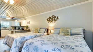 Un dormitorio con dos camas, un ventilador de techo, una cocina al fondo y almohadas decorativas.