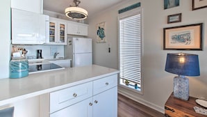 A kitchen with white cabinets, a refrigerator, a coffee maker, and a blue vase. There are framed pictures on the wall and a window with blinds. - Oceanfront Condo w/ Views in North Topsail Beach! (Sneads Ferry)