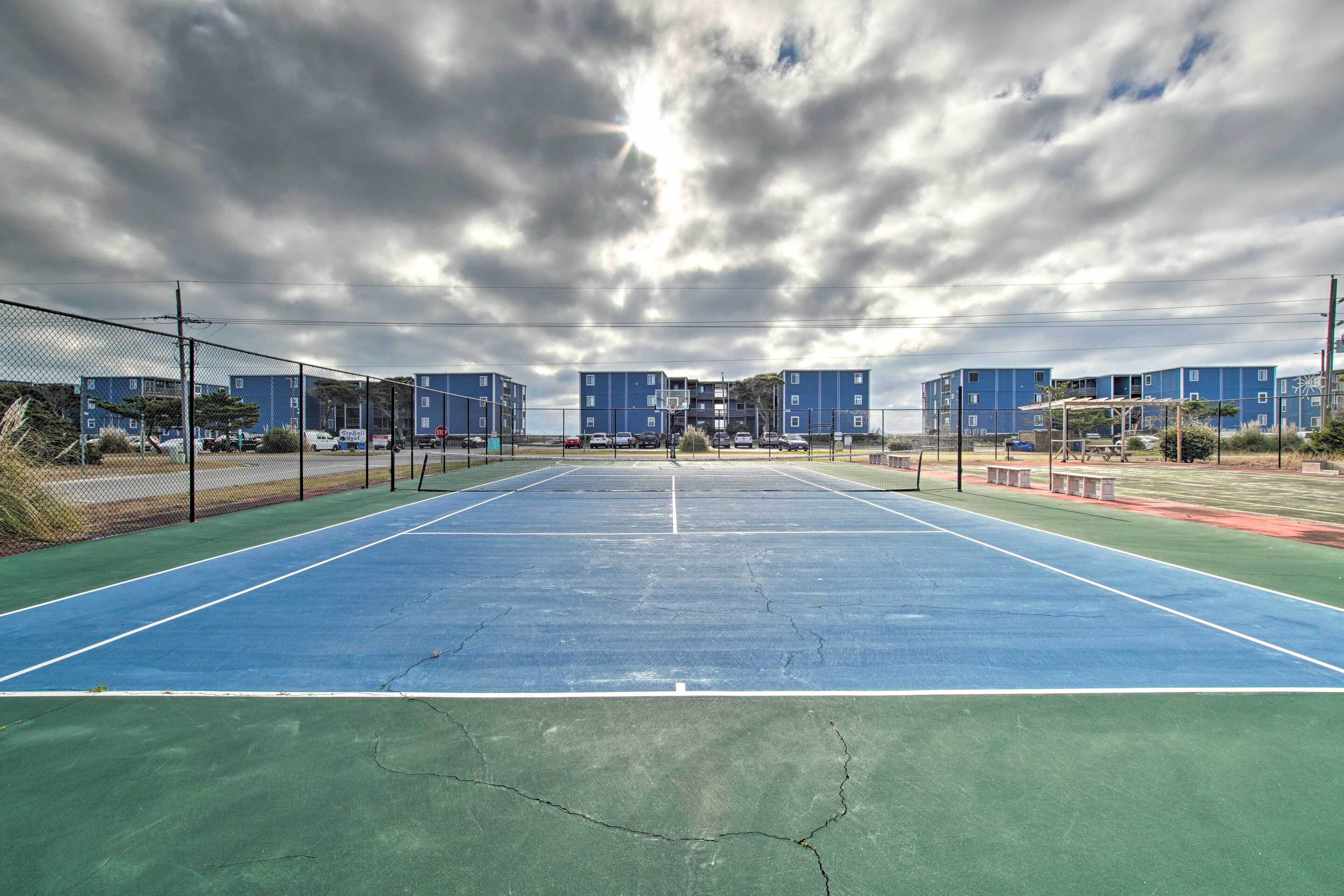 An outdoor tennis court with a net, surrounded by a fence, and buildings in the background.