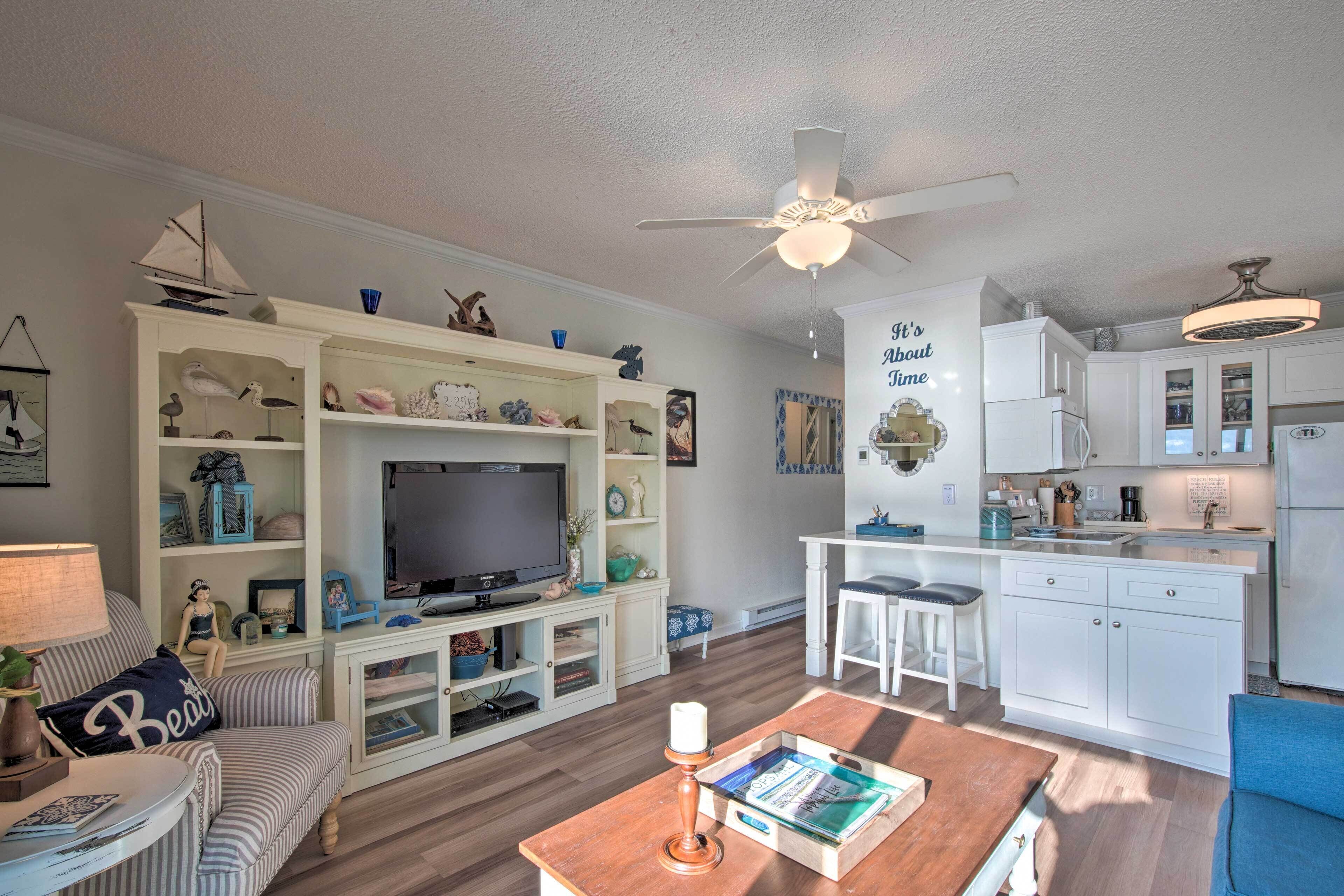 A living room with a TV, a bookshelf, a coffee table, and a kitchen area with white cabinets and a ceiling fan.