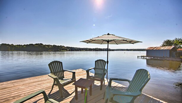 Una terraza de madera con sillas Adirondack y una mesa, con vistas a un lago tranquilo con un muelle y un edificio pequeño al fondo.