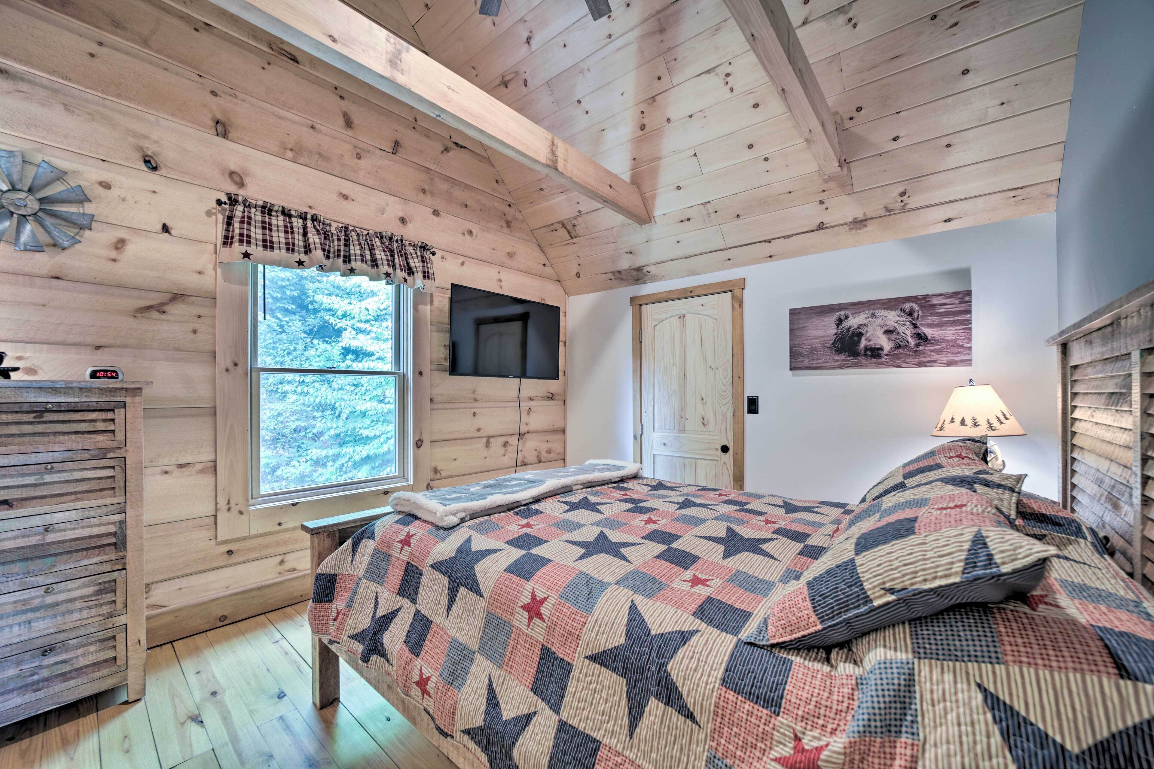 A cozy cabin bedroom with a wooden ceiling, a bed with a star-patterned quilt, a mounted TV, a window with a view of trees, and a door with a bear picture.