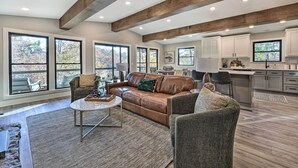 A spacious living room with a brown leather sofa, a round coffee table, and a gray armchair. The room features large windows, a kitchen with white cabinets, and exposed wooden beams on the ceiling. - Hike, Bike & Unwind: Bella Vista Family Home! (Bella Vista)