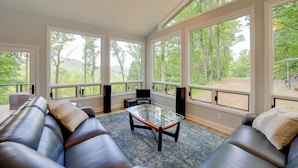 A modern living room with a glass coffee table, leather sofas, and large windows offering a view of a wooded area. - Remote Smoky Mtn Escape w/ Views & Trail Access! (Franklin)
