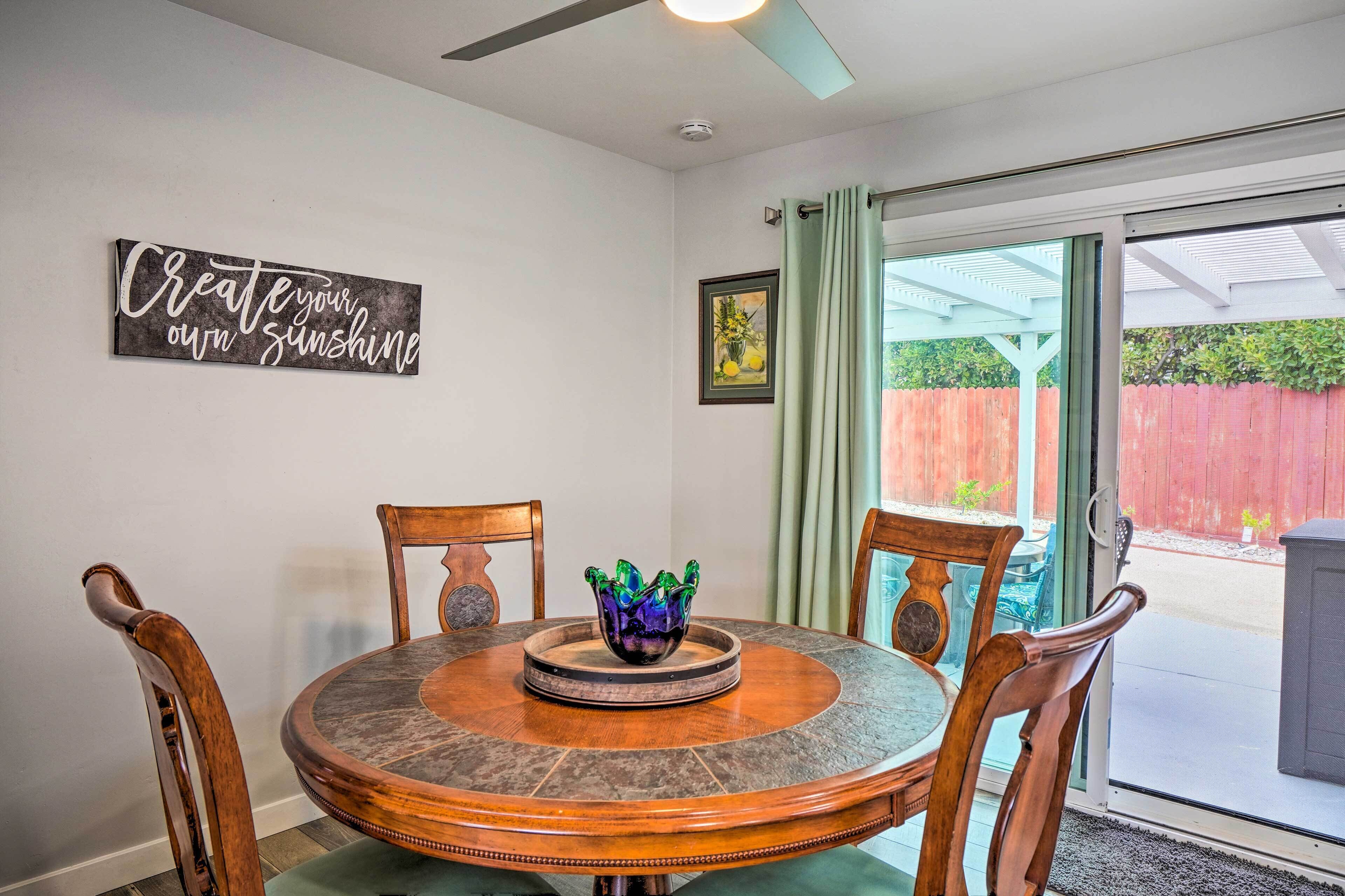 A dining area with a round table, chairs, a decorative bowl, a ceiling fan, and a wall sign.