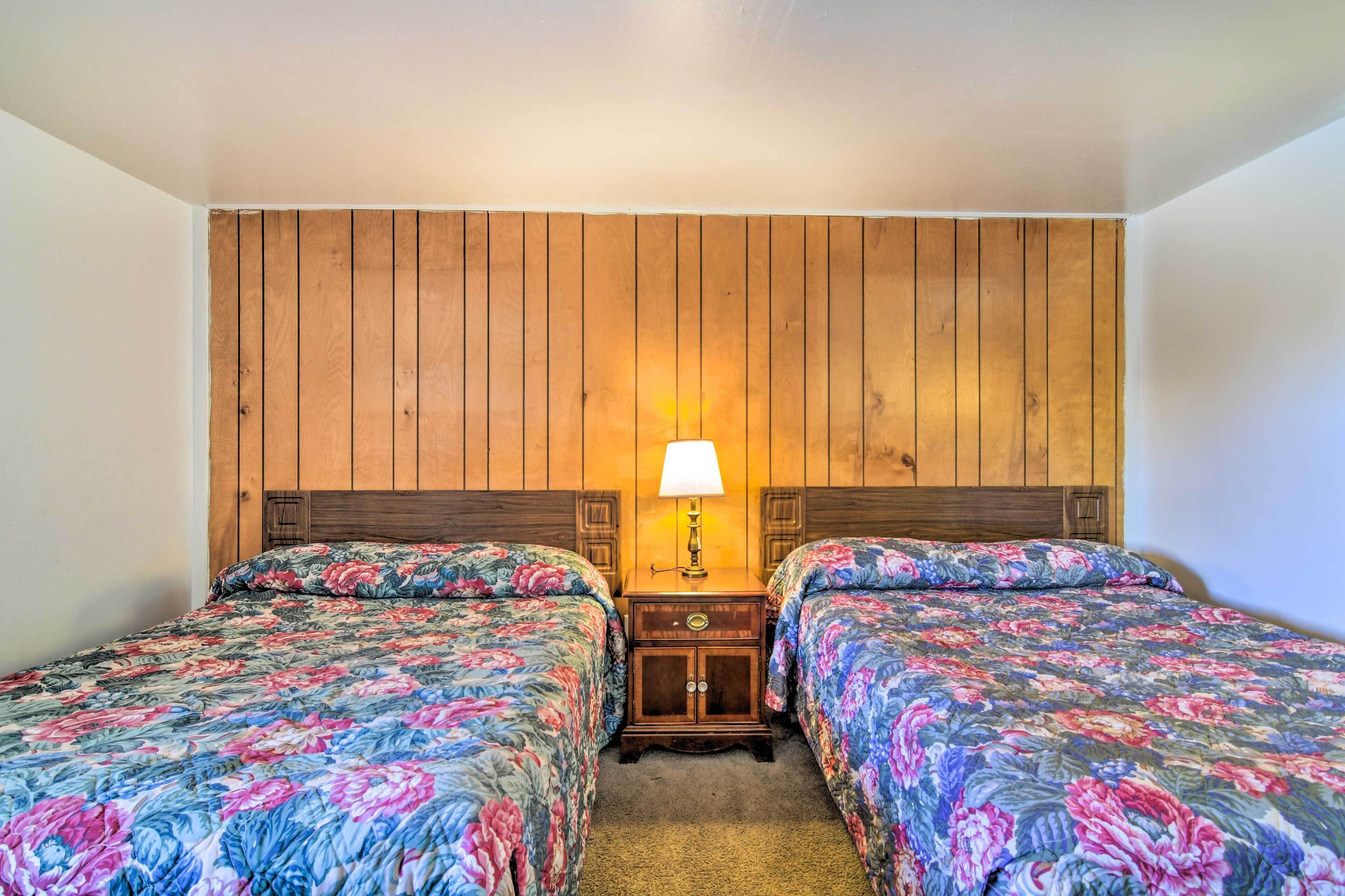 Quarto com duas camas, uma cabeceira de madeira, mesa de cabeceira com abajur e piso coberto por carpete.