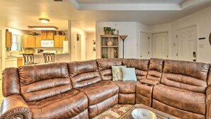 A living room with a brown leather sectional sofa, a kitchen in the background, and a dining area with chairs.