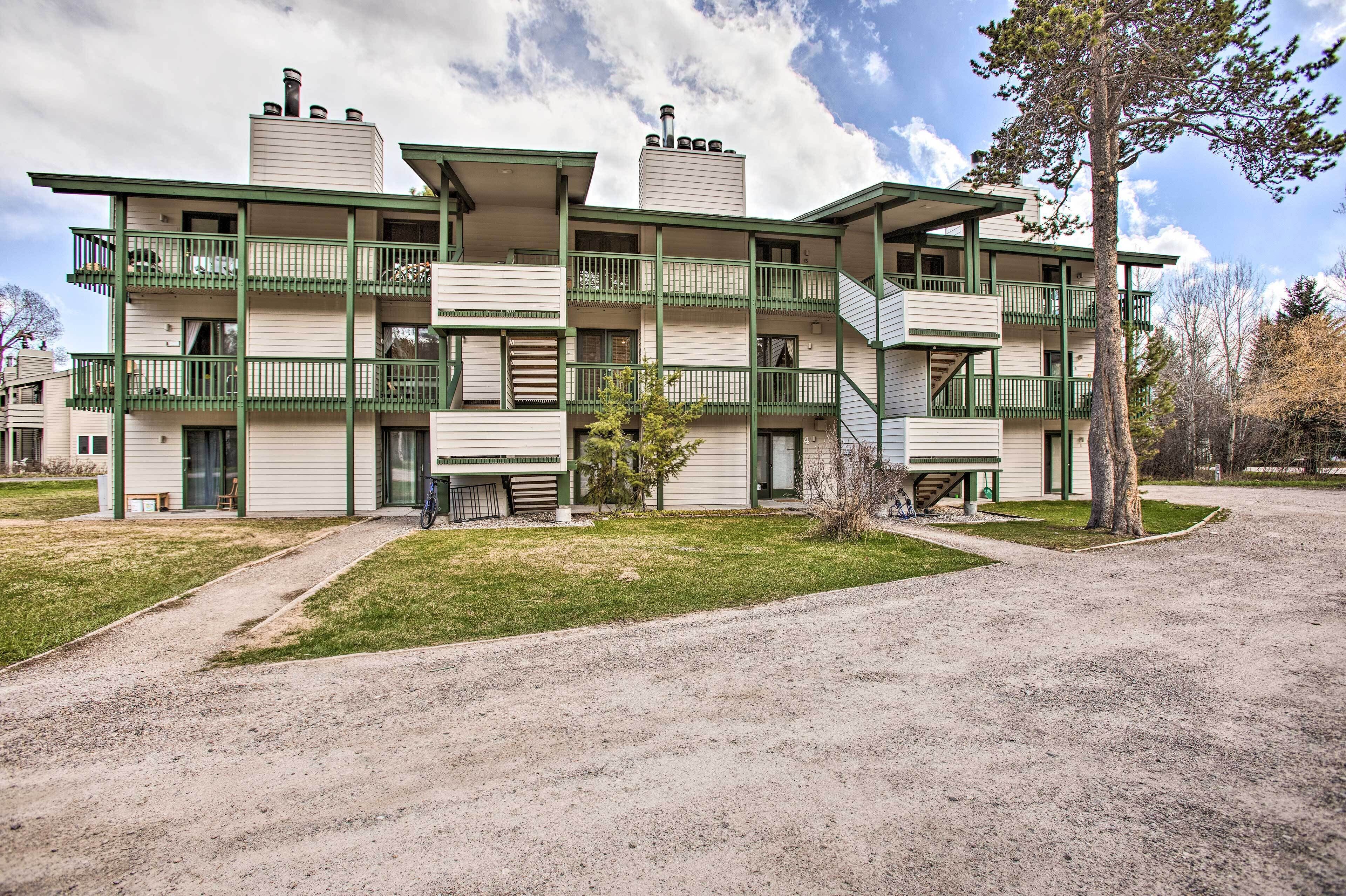 Un edificio de varios pisos con balcones, un espacio al aire libre con sombra para sentarse y un camino de entrada de grava.