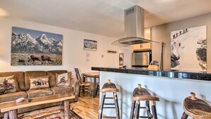 A living room with a sofa, wooden stools, and a kitchen area with a stove and refrigerator. - Skier's Dream: Upscale Condo By Teton Village! (Wilson)