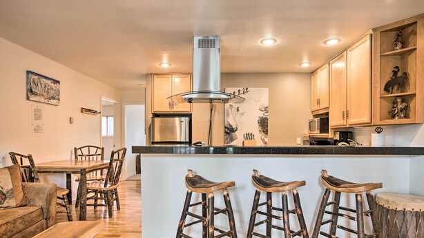 A modern kitchen with wooden cabinets, a stainless steel refrigerator, a central island with stools, and a decorative wall piece. - Skier's Dream: Upscale Condo By Teton Village! (Wilson)