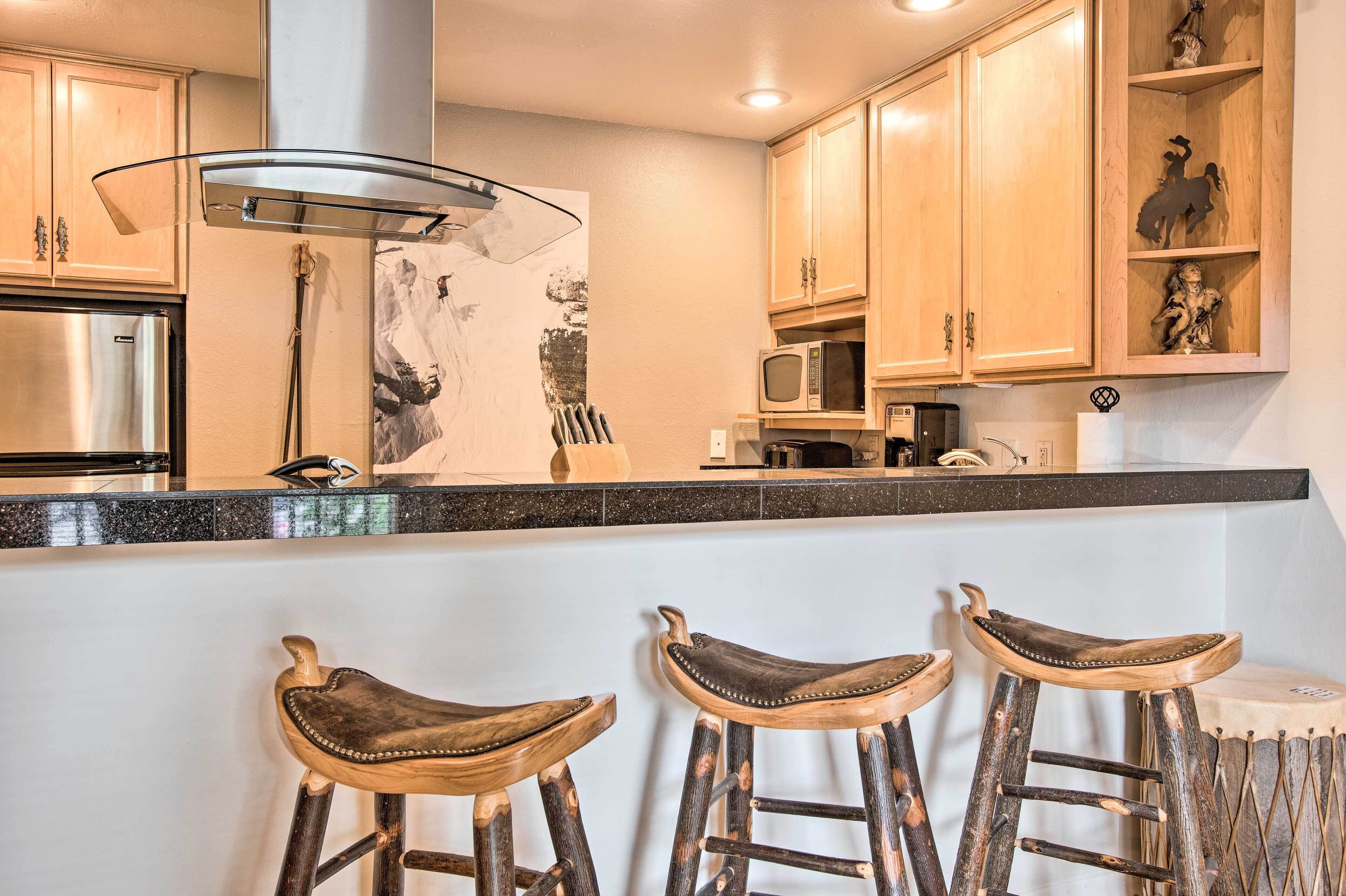 A kitchen with wooden cabinets, a stainless steel refrigerator, a microwave, and a bar area with stools.