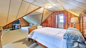 Ein Schlafzimmer mit schrägverdeckter Decke, Holzwänden, einem Bett mit weißer Bettwäsche, einem Nachttisch, einer Lampe und einem Fernseher auf einem Schrank.