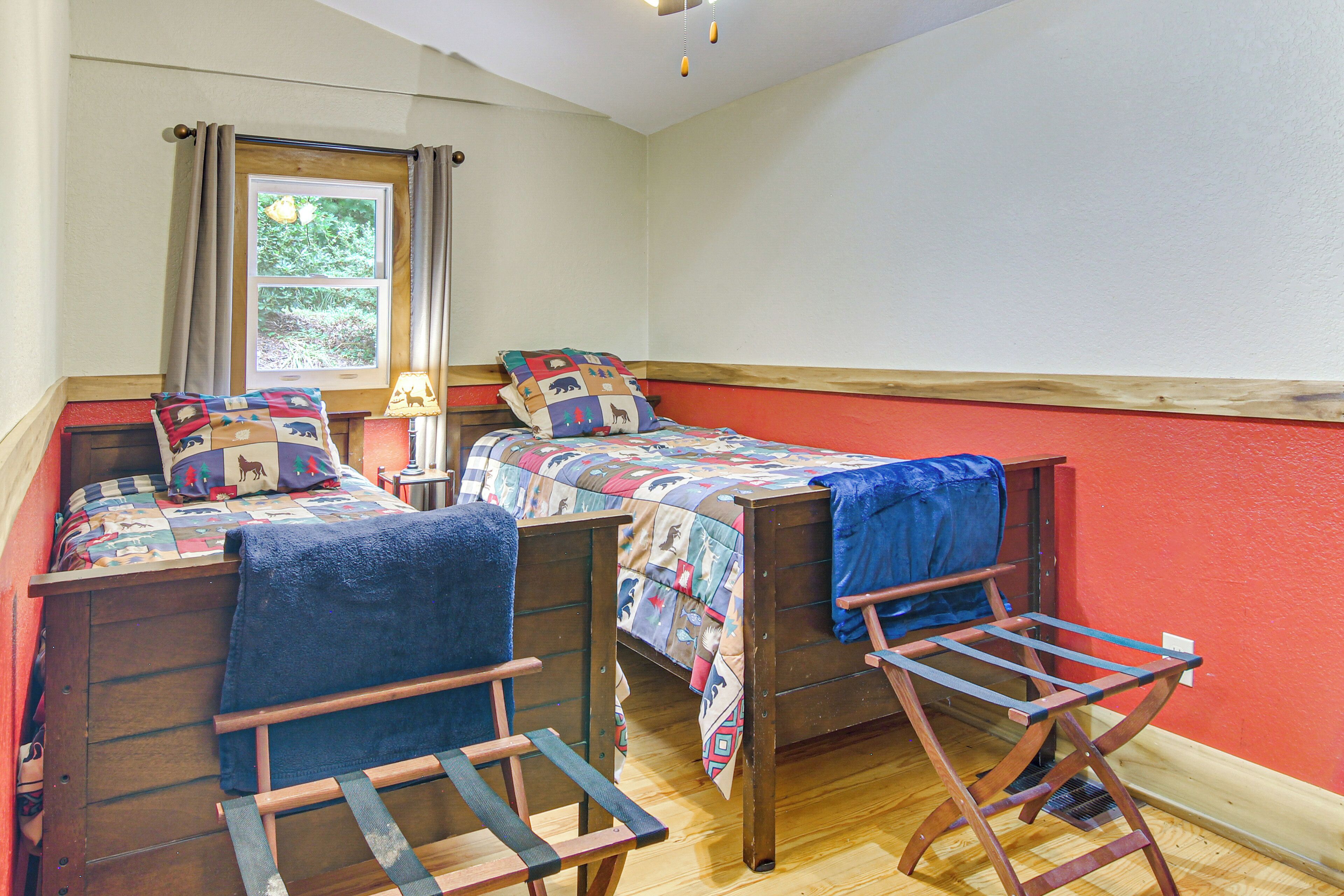 Une pièce avec deux lits en bois, chacun recouvert d’une couverture bleue et d’une courtepointe colorée. On y trouve une fenêtre avec des rideaux, une chaise en bois et une petite table avec une lampe.