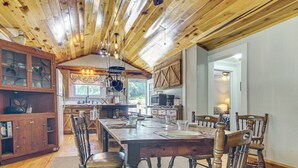 A wooden dining room with a large table set for six, surrounded by chairs. A kitchen with wooden cabinets and a hanging light fixture is visible in the background. - 20-acre Cabin w/ Pond: 6 Mi to River Arts District (Asheville)