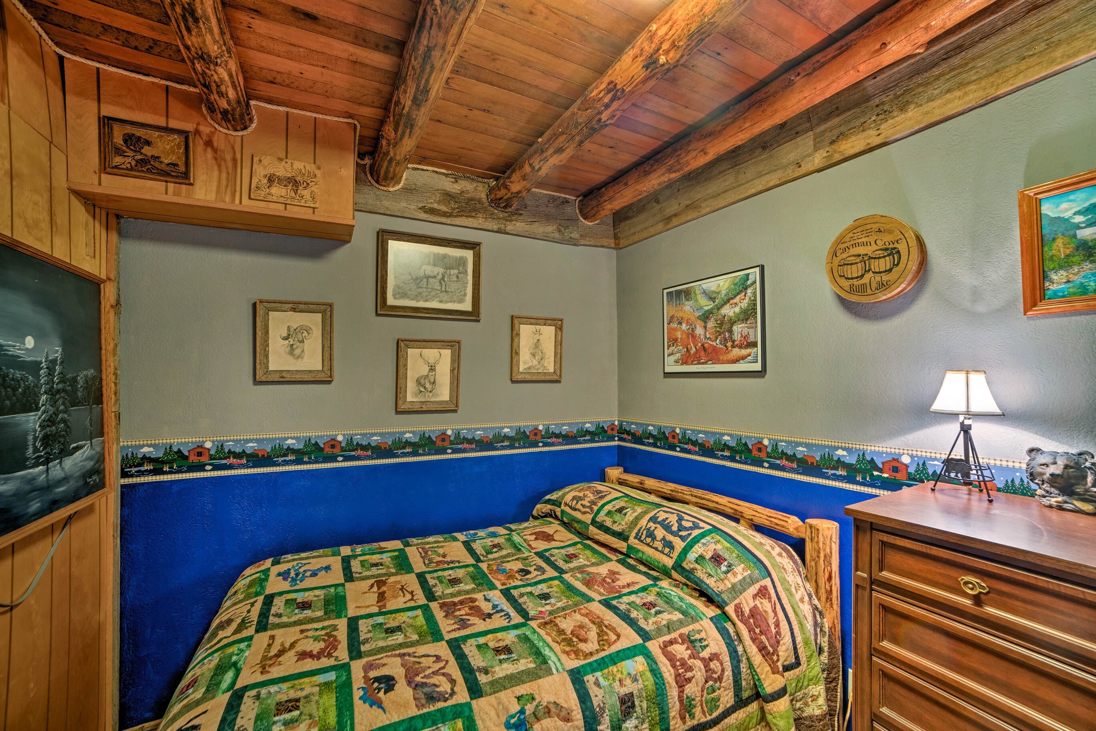 Une chambre avec un mur bleu, des poutres en bois au plafond, un lit recouvert d’une courtepointe, une commode en bois et des tableaux encadrés accrochés au mur.
