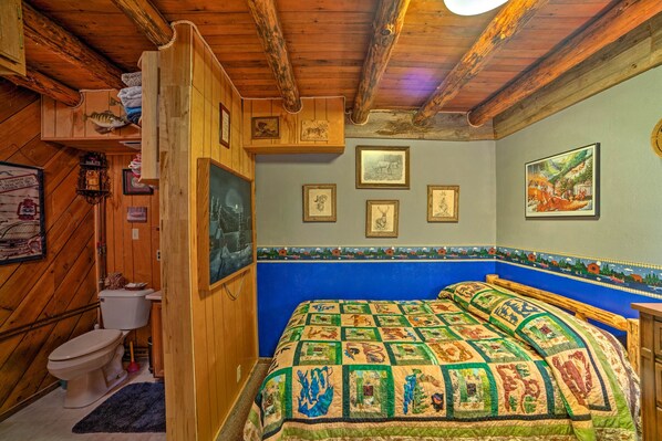 Una habitación rústica con paredes de madera, colcha de patchwork, inodoro y cuadros en la pared.