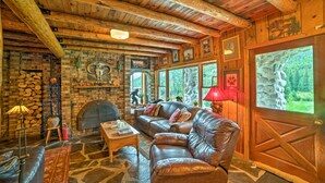 A rustic cabin interior with a stone fireplace, leather sofas, and large windows offering a view of the forest. - 'sasquatch Inn' Retreat Near Fishing & Hunting! (Wise River)