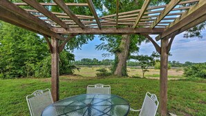 Een terras met een glazen tafel en witte metalen stoelen, beschut onder een houten pergola, omgeven door groen.