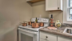 Une cuisine avec des meubles blancs, une cuisinière surmontée de casseroles en cuivre, un évier et une fenêtre donnant sur des arbres.