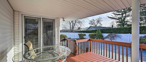 A covered outdoor area with a glass table and chairs, a wooden deck, and a view of a residential area with a white fence and trees.