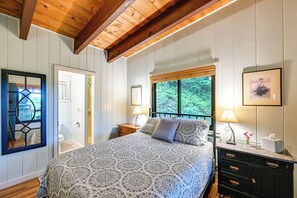 A bedroom with a large bed, wooden ceiling beams, a window with a view of trees, a bedside table with a lamp, and a dresser. - Sweet Birch' Bryson City Cottage w/ Views (Bryson City)