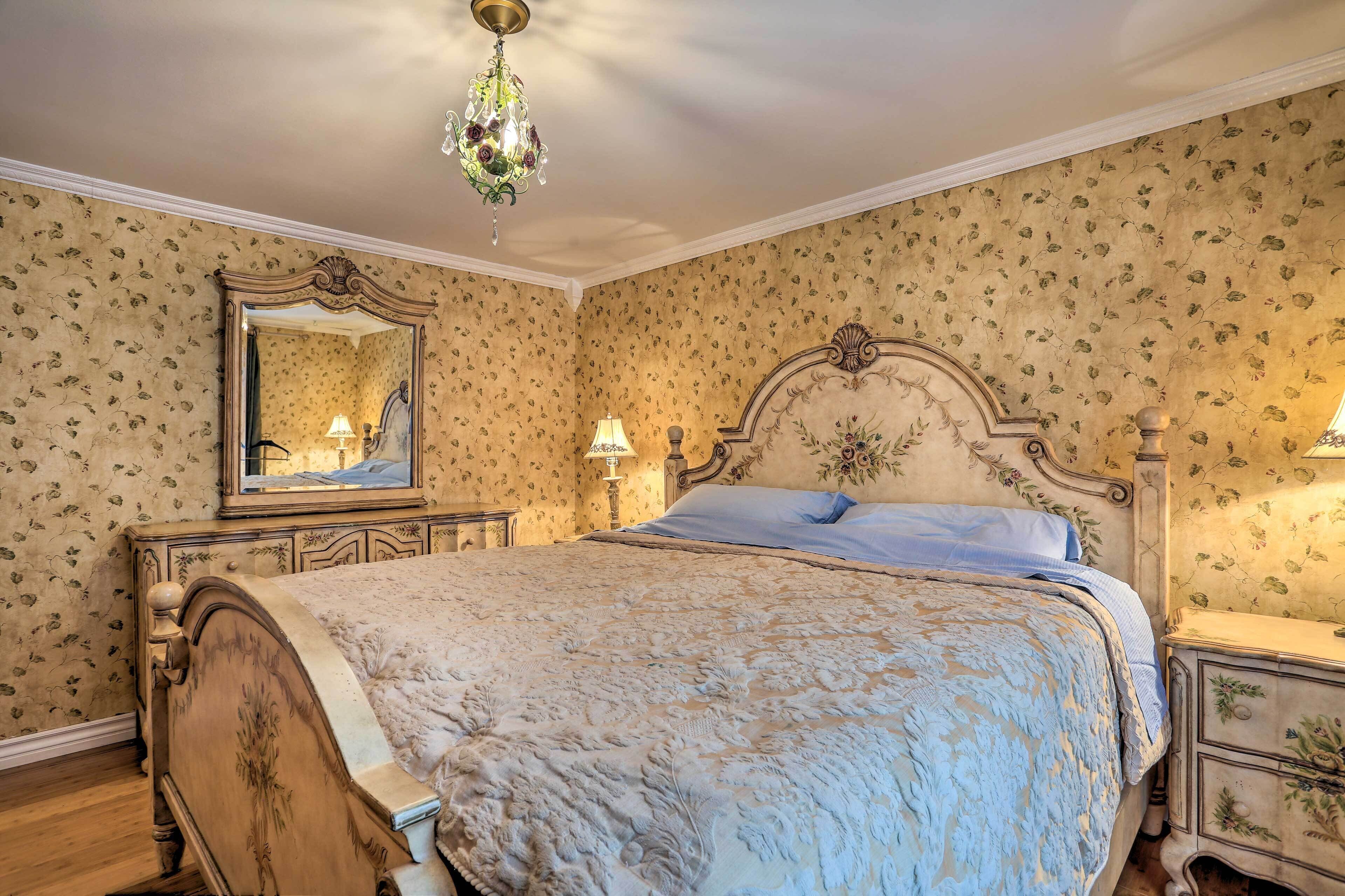 A bedroom with a large bed, ornate headboard, floral wallpaper, and a chandelier.