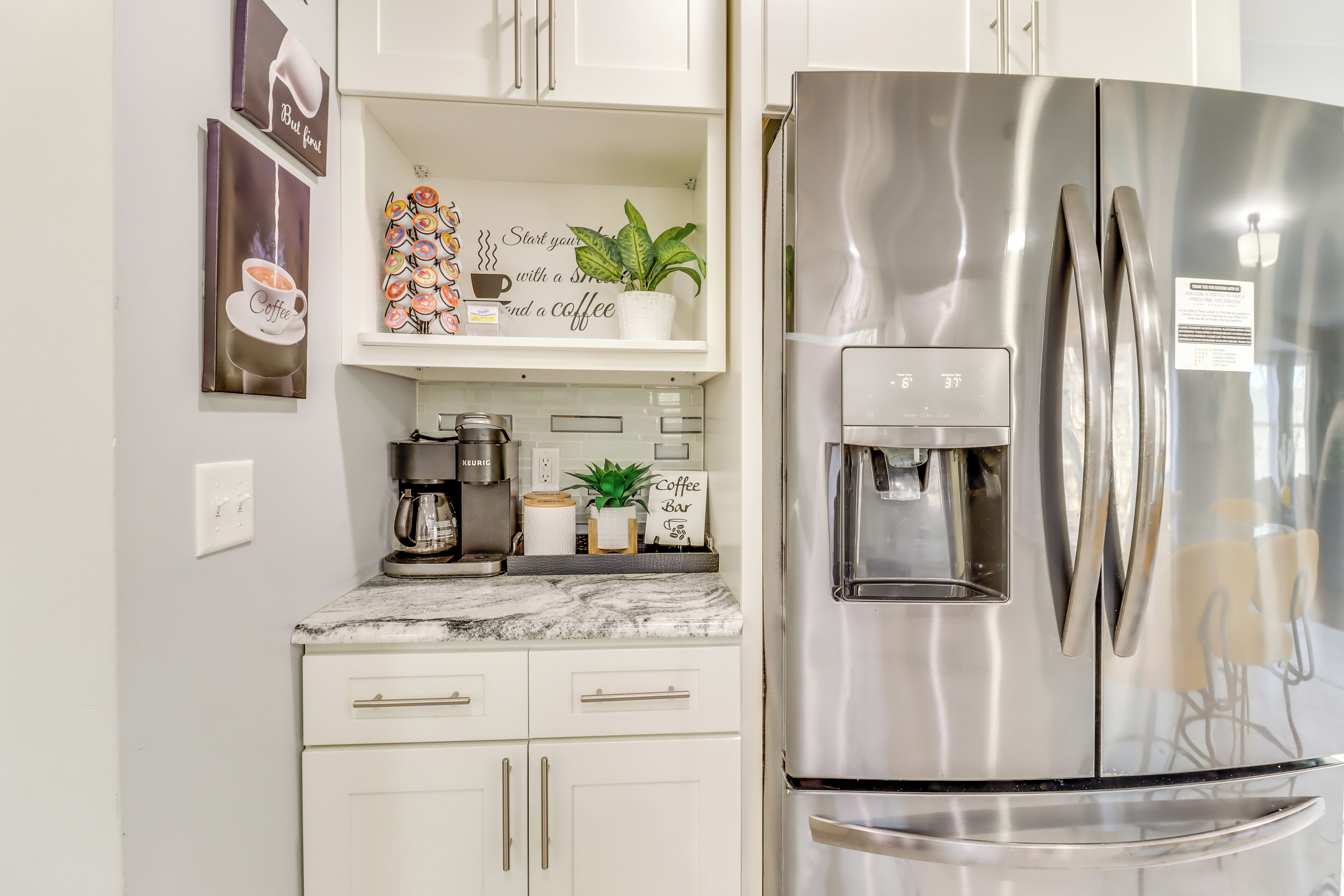 Una cocina con armarios blancos, un refrigerador de acero inoxidable, una cafetera y una repisa con adornos relacionados con el café.