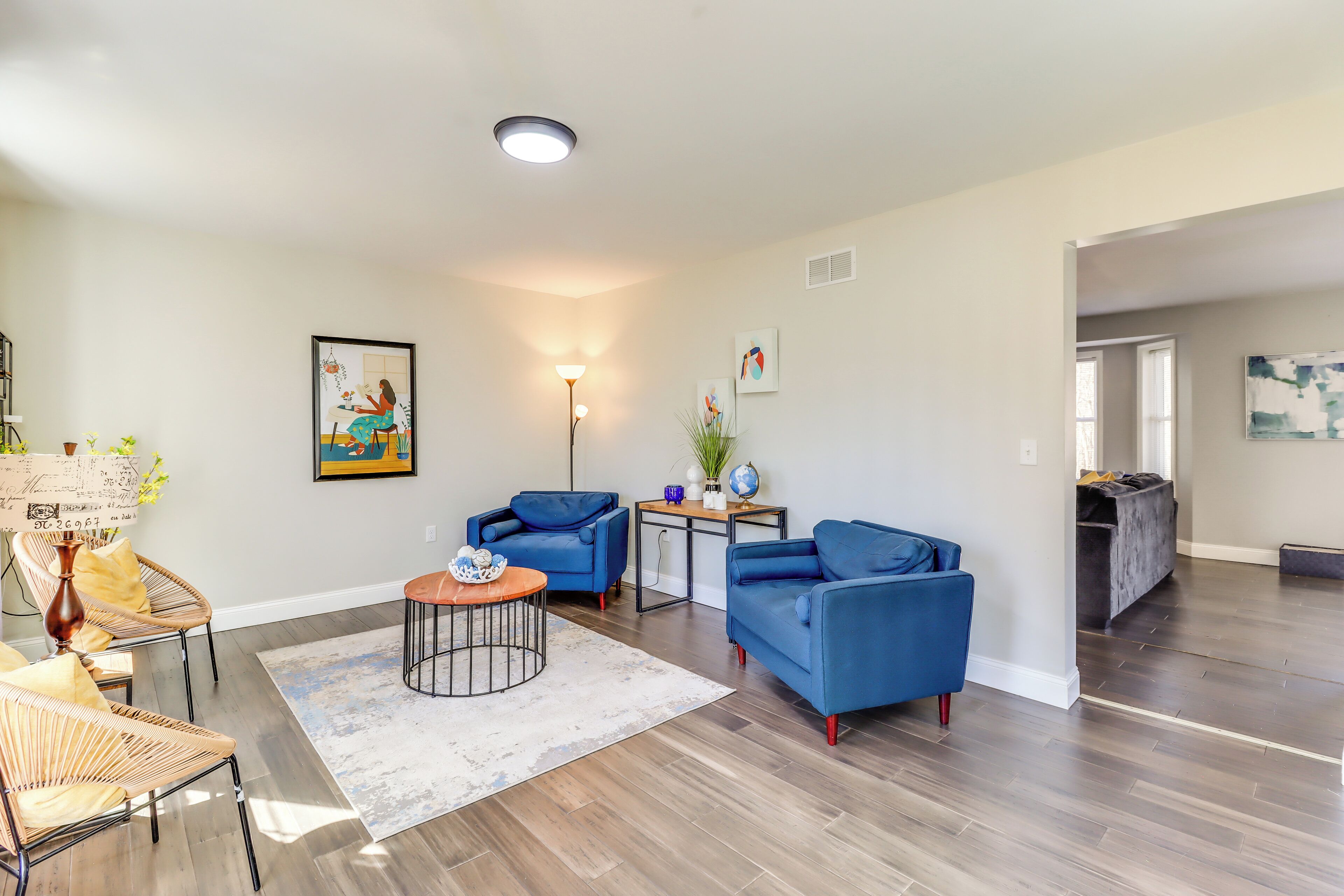 A spacious living room with hardwood flooring, a blue sofa, a coffee table, and a chair. There is a painting on the wall and a lamp on a side table.