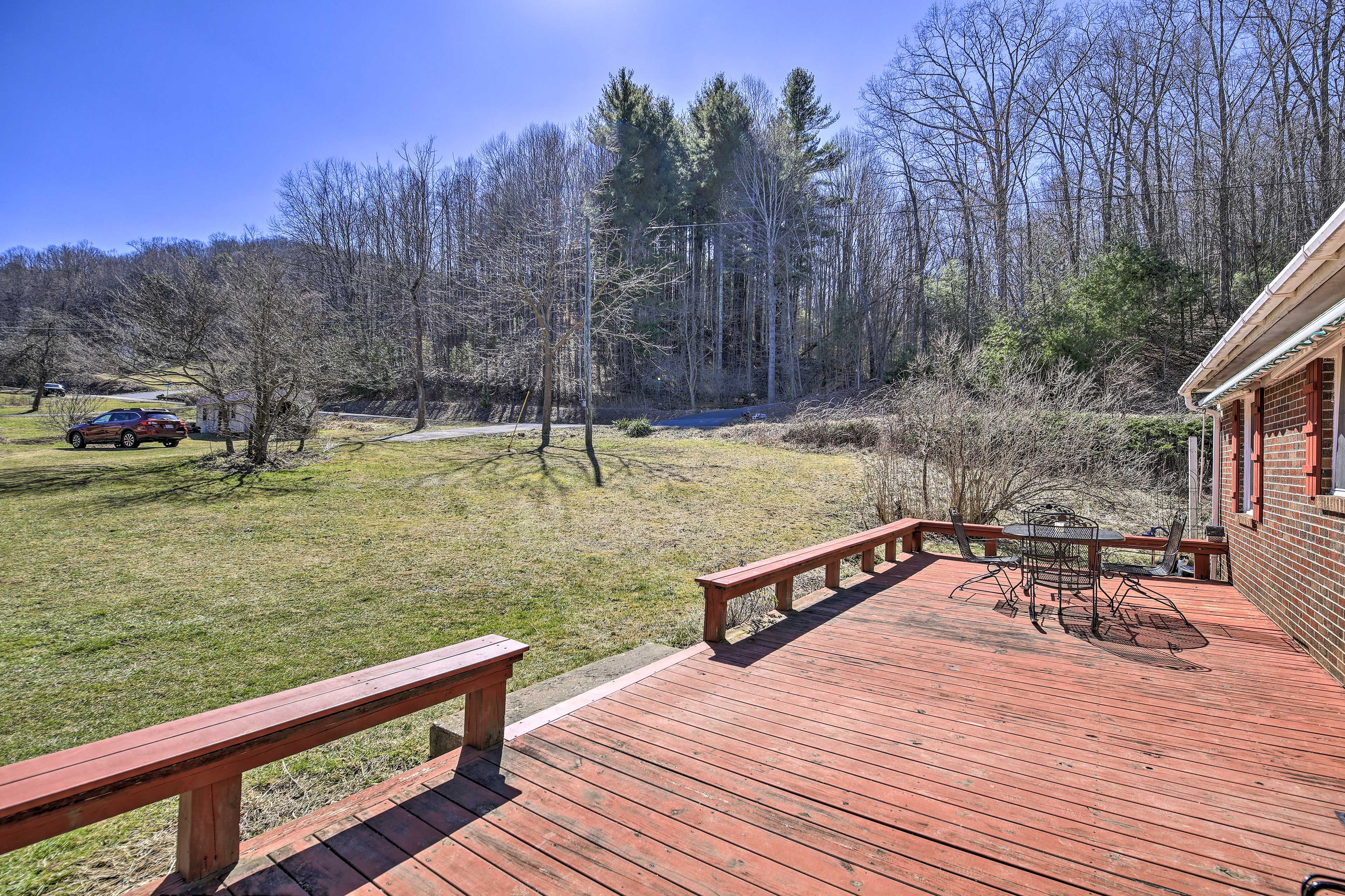 A wooden deck with a table and chairs, overlooking a grassy area with trees and a parked car.