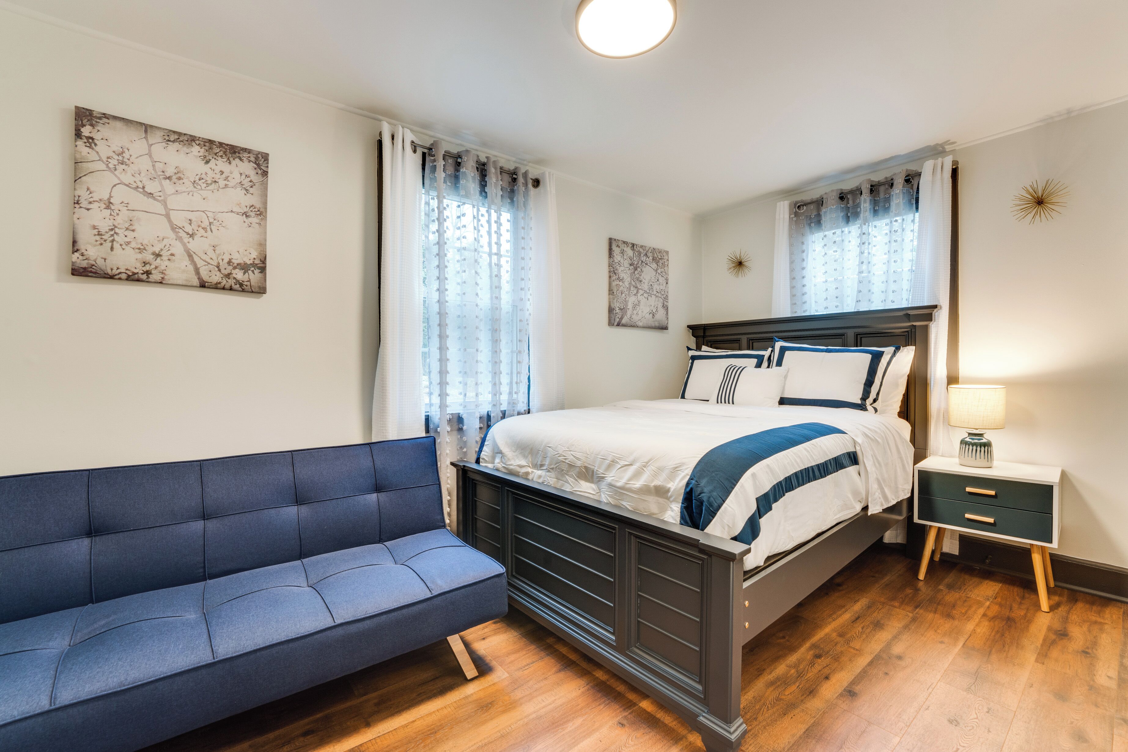 A bedroom with a bed, a blue sofa, a bedside table with a lamp, and two windows with curtains.