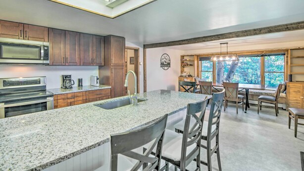 A modern kitchen with granite countertops, stainless steel appliances, and wooden cabinets. There is a large island with bar stools, and a dining area with a view of trees through large windows. - 'moose Park Lodge' Charming Walden Retreat! (Walden)
