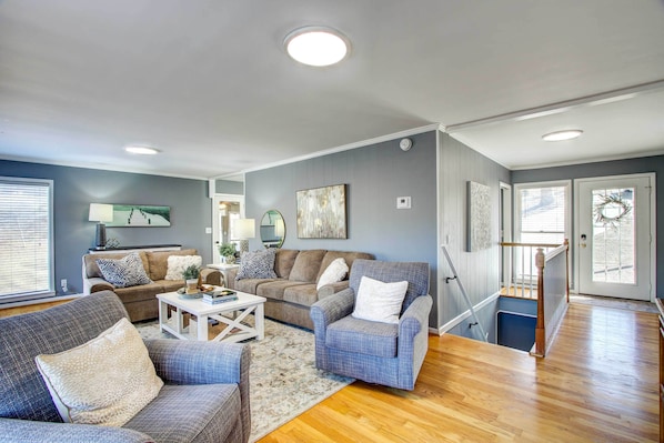 A living room with a grey couch, a blue armchair, and a coffee table. There is a staircase leading to a balcony with a railing. - Waterfront Florence Home w/ Private Dock! (Florence)