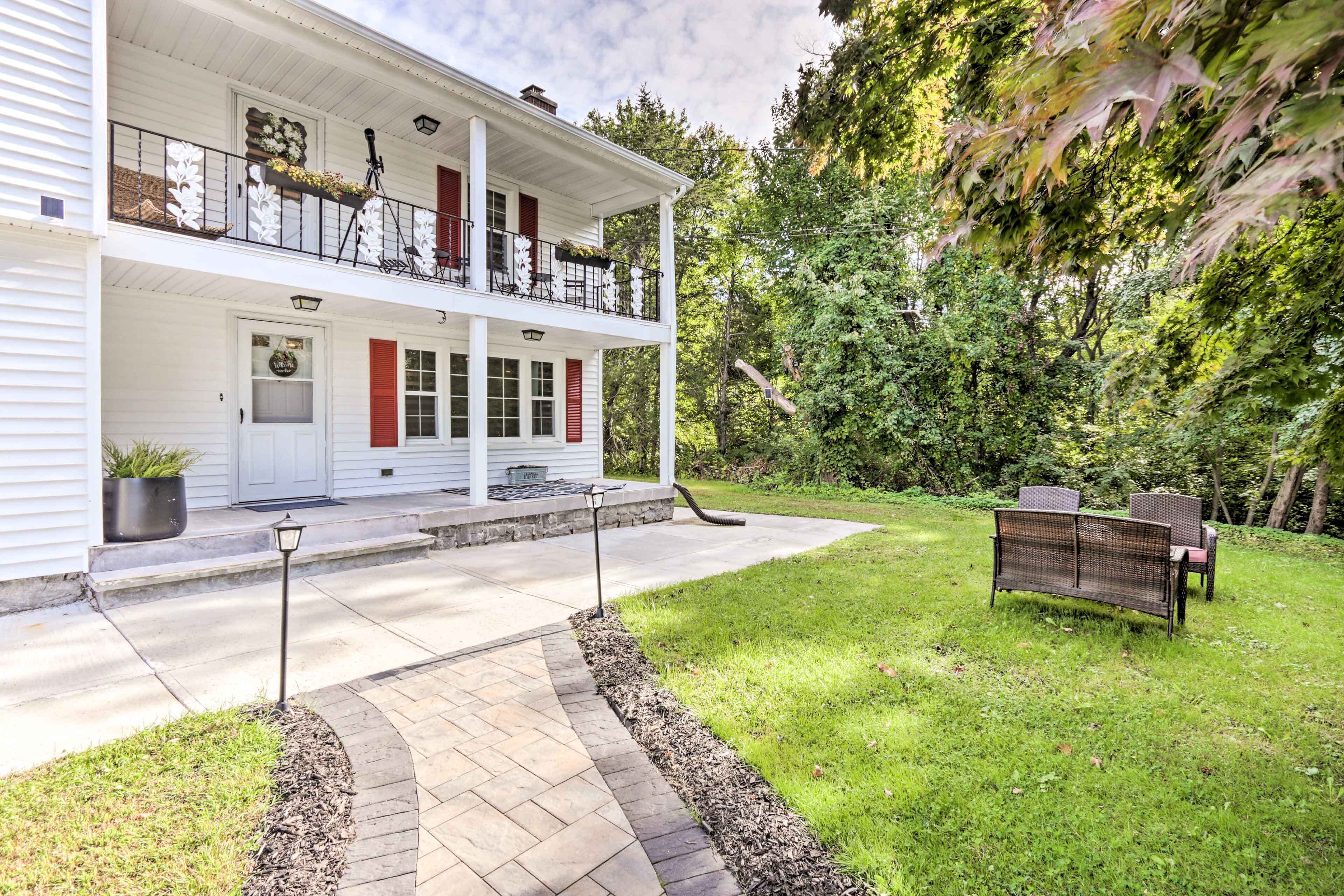 A two-story house with a balcony, a front door, and a pathway leading to a grassy area with outdoor furniture.