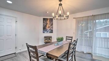 Uma sala de jantar com mesa de madeira, cadeiras, um candelabro, um quadro na parede e uma janela com cortinas leves.