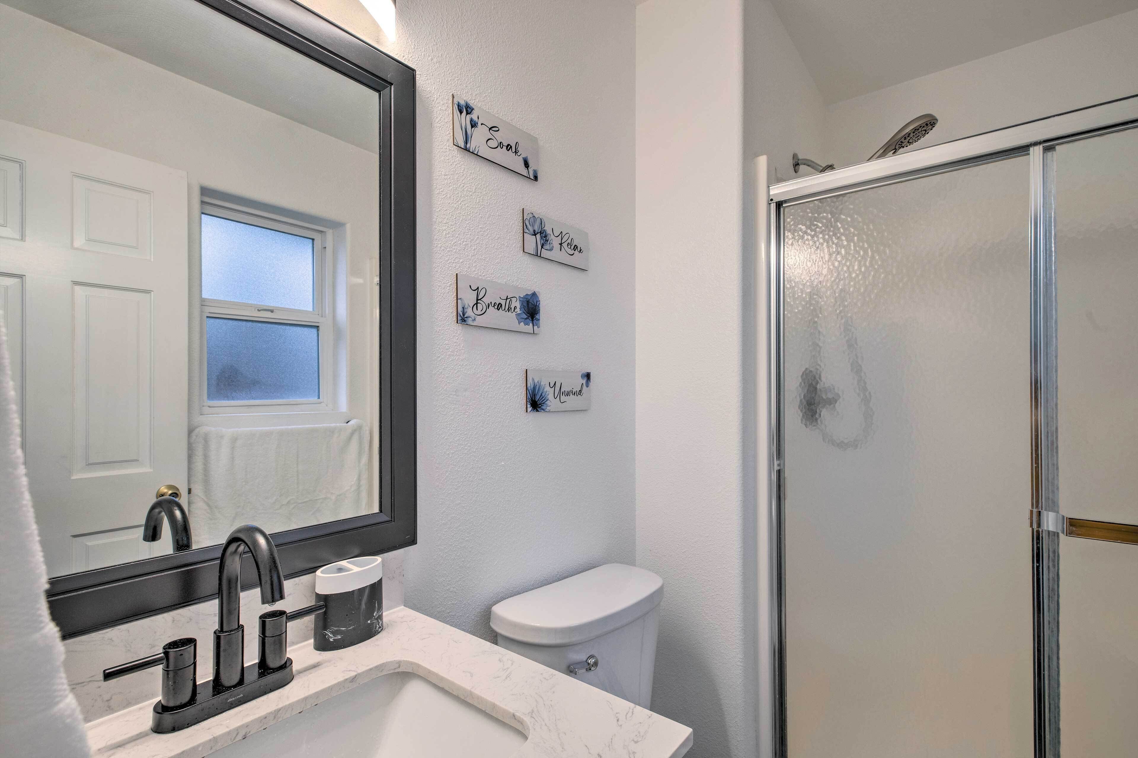 Un baño con lavamanos blanco, grifo negro, un espejo, inodoro y mampara de cristal para la ducha.