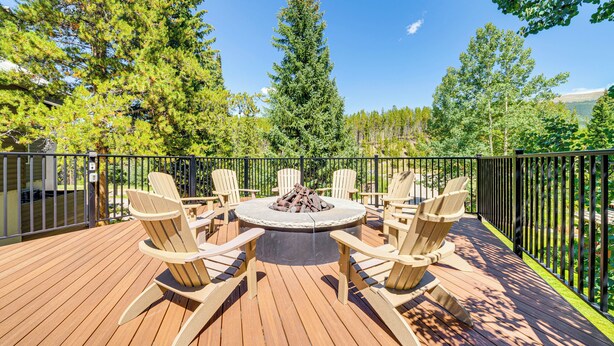 A deck with a circular fire pit surrounded by six Adirondack chairs, overlooking a forested area.