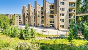 A multi-story residential building with balconies, a covered outdoor area, and a natural surrounding with trees and grass.