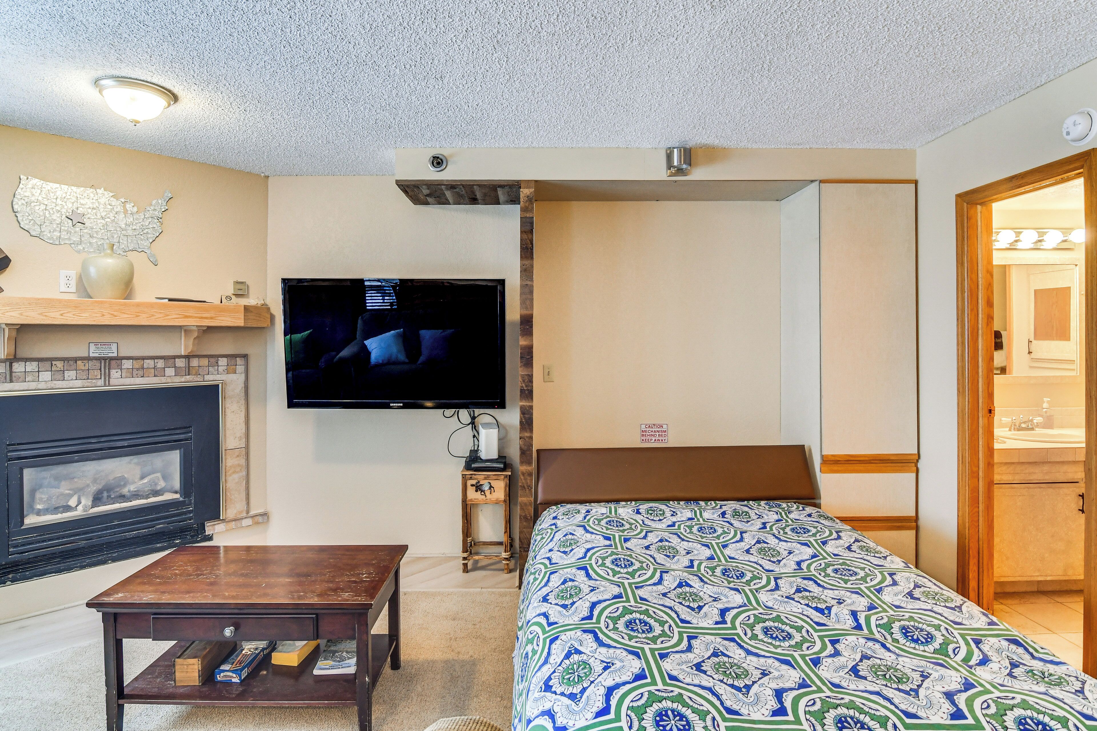 A hotel room with a fireplace, a television mounted on the wall, a bed with a patterned bedspread, a wooden coffee table, and a small side table with a lamp.