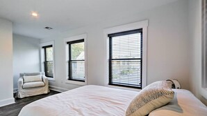 A bedroom with a large bed, two windows with blinds, a chair, and a small table. - Updated Downtown Red Bank Home 5 Mi to Beach! (Red Bank)