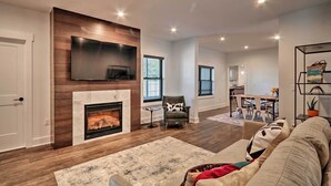 A living room with a fireplace, a TV mounted on the wall, a dining area with a table and chairs, and a kitchen in the background. - Updated Downtown Red Bank Home 5 Mi to Beach! (Red Bank)