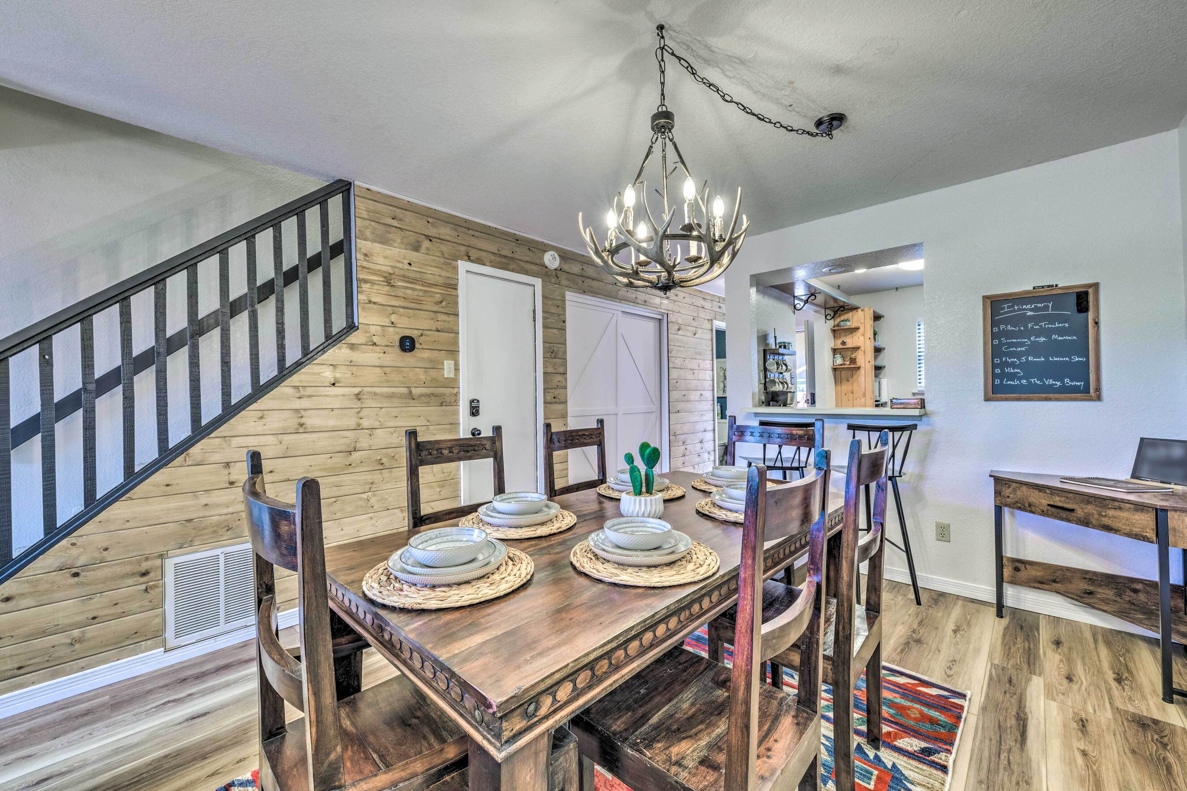 A dining area with a wooden table set for four, surrounded by chairs. A chandelier hangs from the ceiling, and a chalkboard menu is visible on the wall.