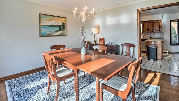 Un comedor con una mesa y sillas de madera, una araña, un cuadro de un paisaje montañoso y vista a la cocina.