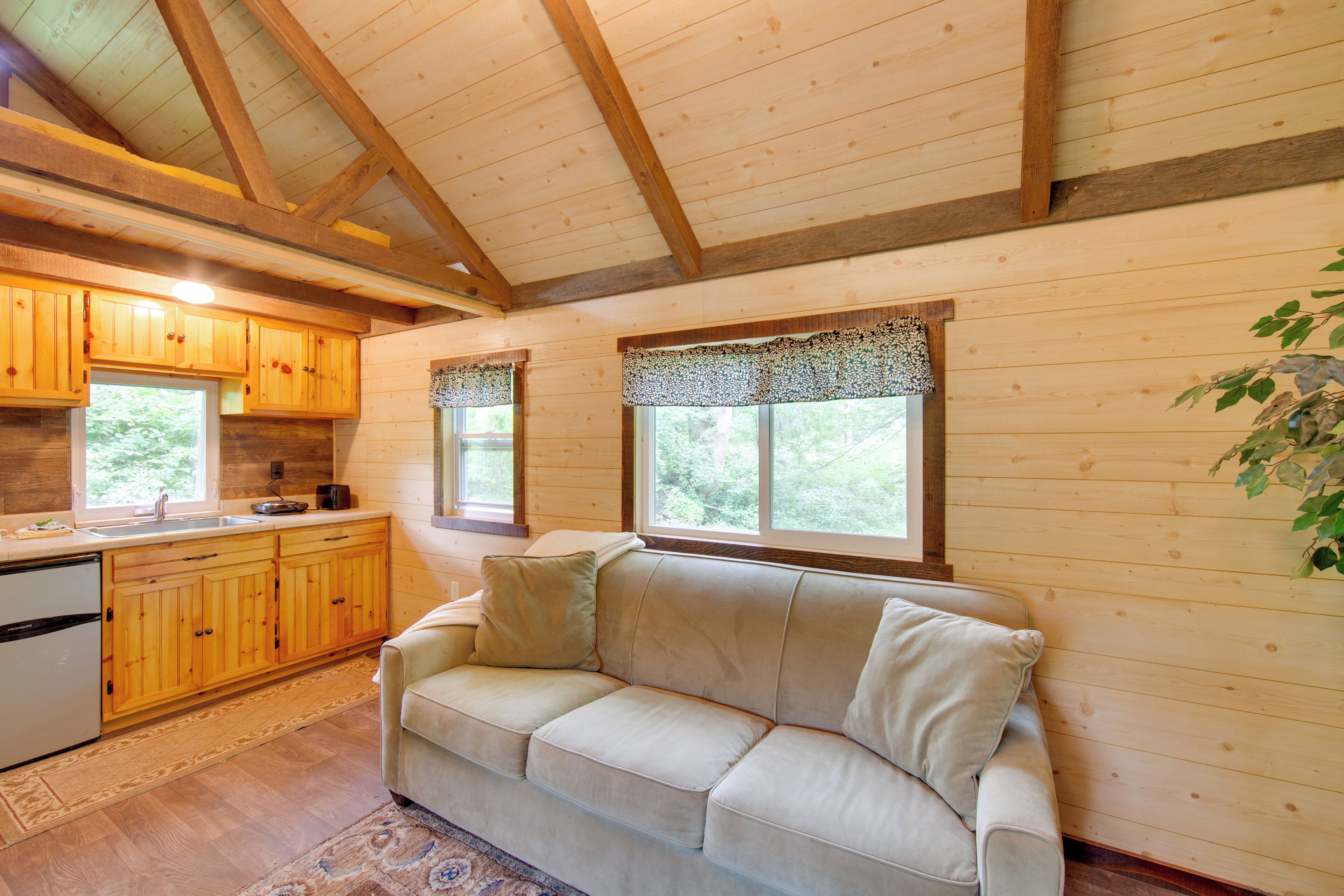 A cozy cabin interior with wooden walls, a beige sofa, a kitchen with wooden cabinets, and a window with a decorative valance.