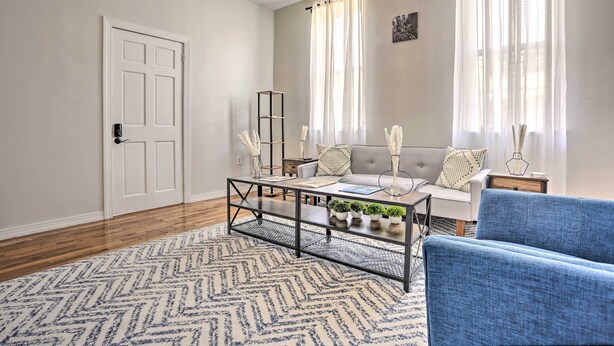 Sala de estar con sofá, una mesita de centro y un sillón azul. Se ve una puerta y una ventana con cortinas.