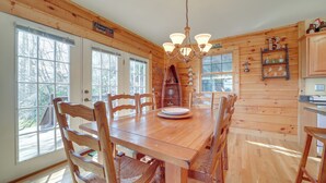 Una mesa de comedor de madera con sillas, una araña y una estantería en una habitación de estilo rústico con ventanas amplias.