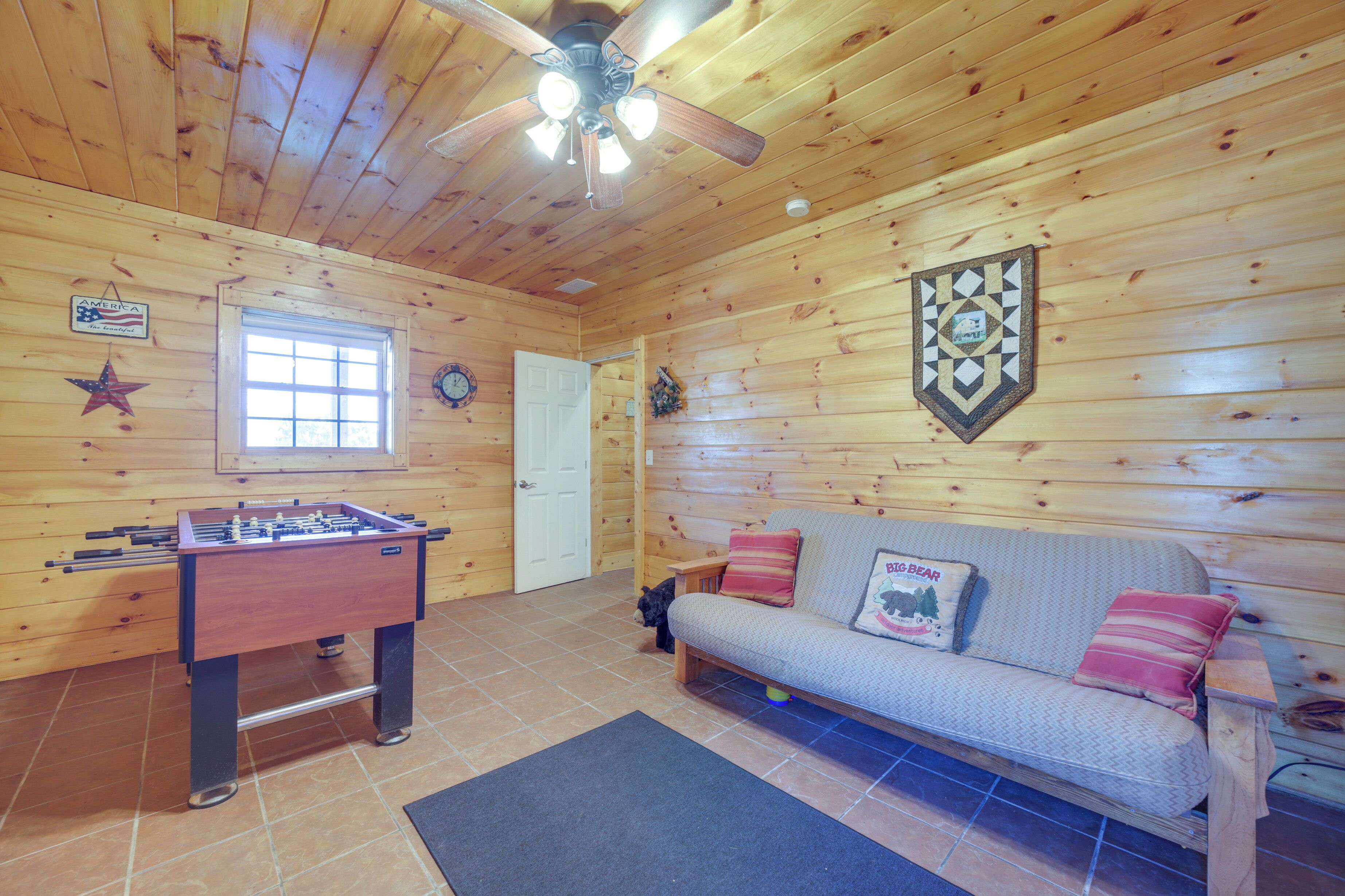 Una habitación acogedora con paredes de madera, una mesa de fútbolín, un sofá con almohadones y un ventilador de techo, además de una ventana.