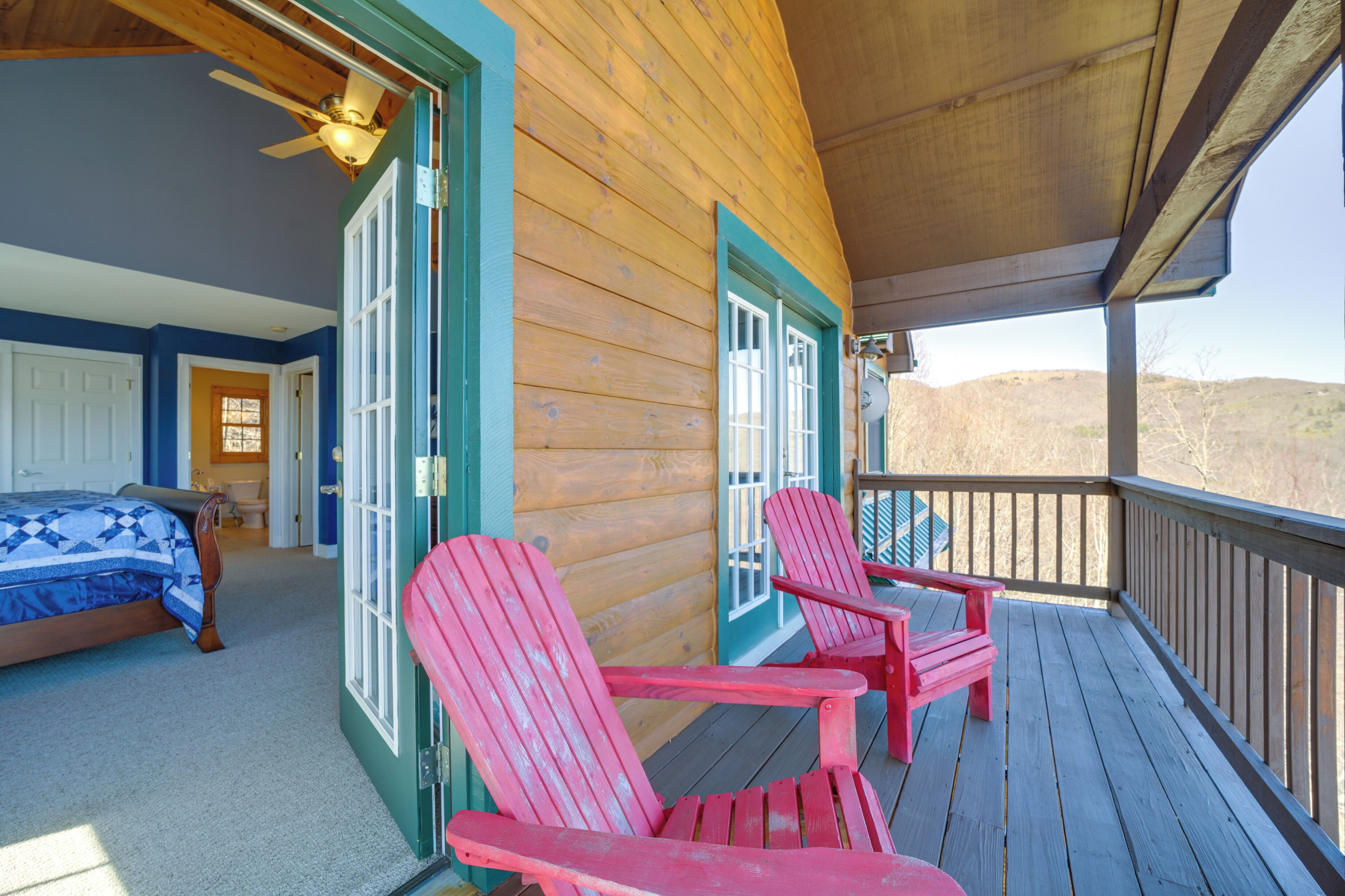 A wooden cabin with a bedroom, two Adirondack chairs, and a balcony overlooking a hilly landscape.